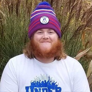 Photo of Dan Gallatin smiling at the camera. They have long red hair and a long beard. They are wearing a purple, red, white, and blue beanie.