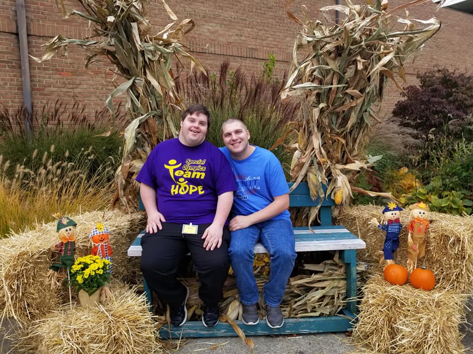Two people sitting on a bench smiling at the camera. They have hay bales around them covered in fall décor.