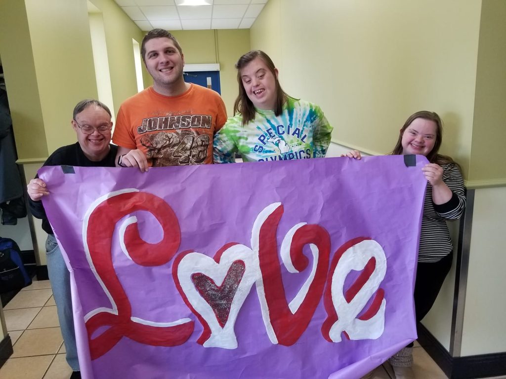 Group of four people smiling and holding a large purple banner that has the word love painted on it with red and white.