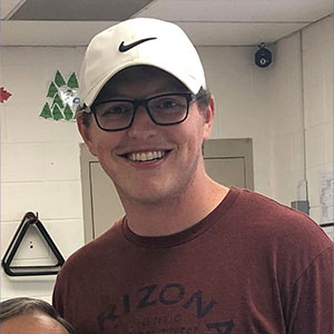 Photo of Logan Milligan smiling at the camera. Wearing a white Nike hat, Black rimmed glasses, and a red t-shirt.